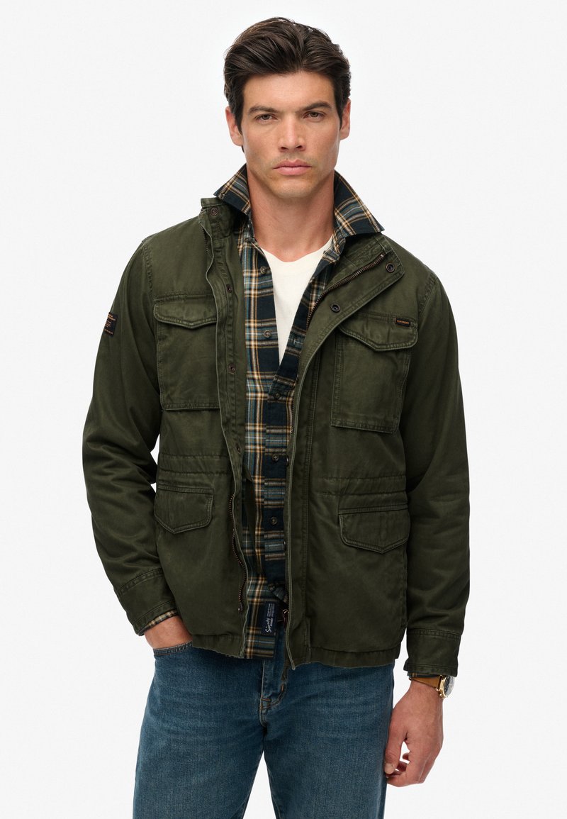 Veste militaire vert olive avec quatre poches à l'avant, superposée sur une chemise à carreaux bleu marine et jaune, un jeans en denim et un t-shirt blanc.