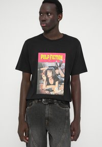 Νεαρός άνδρας που φοράει μαύρο μπλουζάκι με γραφικά από το "Pulp Fiction" και σκούρα τζιν, στέκεται με τα χέρια στις πλευρές, κοιτάζοντας ελαφρώς αριστερά.