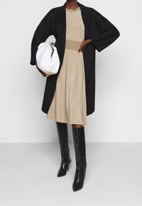 Manteau long noir, robe beige avec taille côtelée, et un sac à main blanc. La mannequin porte des bottes noires ajustées jusqu'aux genoux, mettant en valeur une texture lisse et une forme élégante.