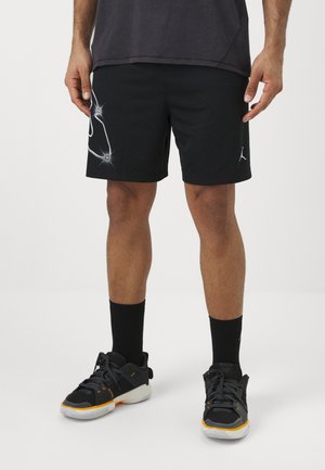 Mężczyzna w czarnych krótkich spodenkach sportowych z białym wzorem na lewym udzie, czarnych skarpetach i czarnych trampkach z białą i pomarańczową podeszwą.