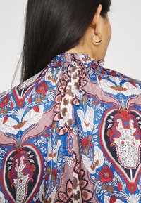 Blusa multicolore con pattern floreale e paisley in blu, rosa e bianco, con scollatura arricciata e chiusura a pulsante sul retro.