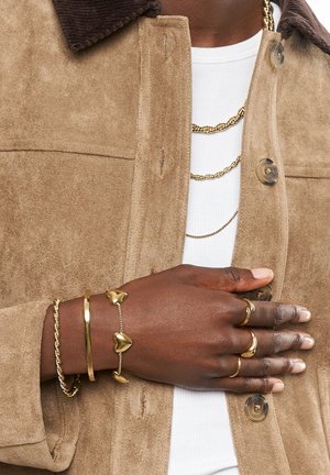 Veste en daim beige avec un col en velours côtelé marron, portée par-dessus une chemise blanche. Main ornée de plusieurs bagues et bracelets en or, incluant des formes en cœur.