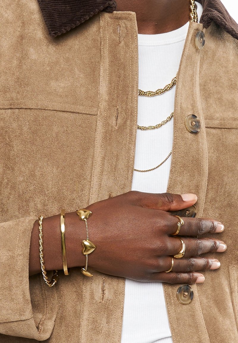 Beige Wildlederjacke mit einem braunen Cordkragen, über einem weißen Hemd getragen. Hand geschmückt mit mehreren goldenen Ringen und Armbändern, darunter herzförmige.