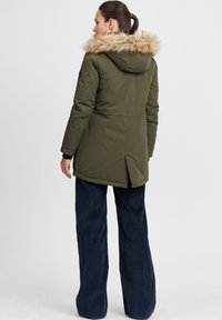 Olive green parka with a faux fur hood, featuring a zip closure and a high back slit, paired with navy corduroy wide-leg pants.