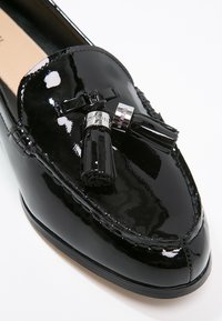 Mocassin en cuir verni noir avec des glands décoratifs et des accents en métal argenté sur un fond blanc.