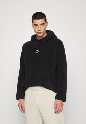 Černá fleecová mikina s kapucí a přední kapsou, se stahovací šňůrkou a bílým logem. Má uvolněný střih a strukturovanou tkaninu. Kombinována se světlými kalhotami.