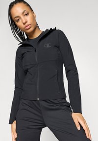 Melnā zipa jakā ar pieguļošu dizainu, ar augstu apkakli, kabatām ar pārliku un smalku logotipu uz krūtīm. Mīksts, elastīgs audums.