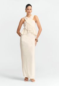 Robe longue de couleur crème en tissu léger. Présente une couche froncée et un design de bretelles croisées. Portée avec des accessoires en or massif.