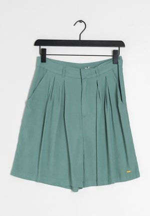 Shorts taille haute plissés couleur sarcelle avec passants de ceinture et poches latérales suspendus sur un cintre noir sur fond blanc.