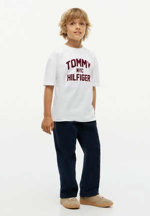 Blond jongetje staat, draagt een wit Tommy Hilfiger T-shirt, donkere broek en beige schoenen tegen een effen lichte achtergrond.
