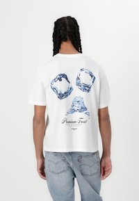 T-shirt bianco a maniche corte con grafiche di cubetti di ghiaccio blu e disegni di gocce d'acqua sul retro, con il testo "Premium Frost" sotto.
