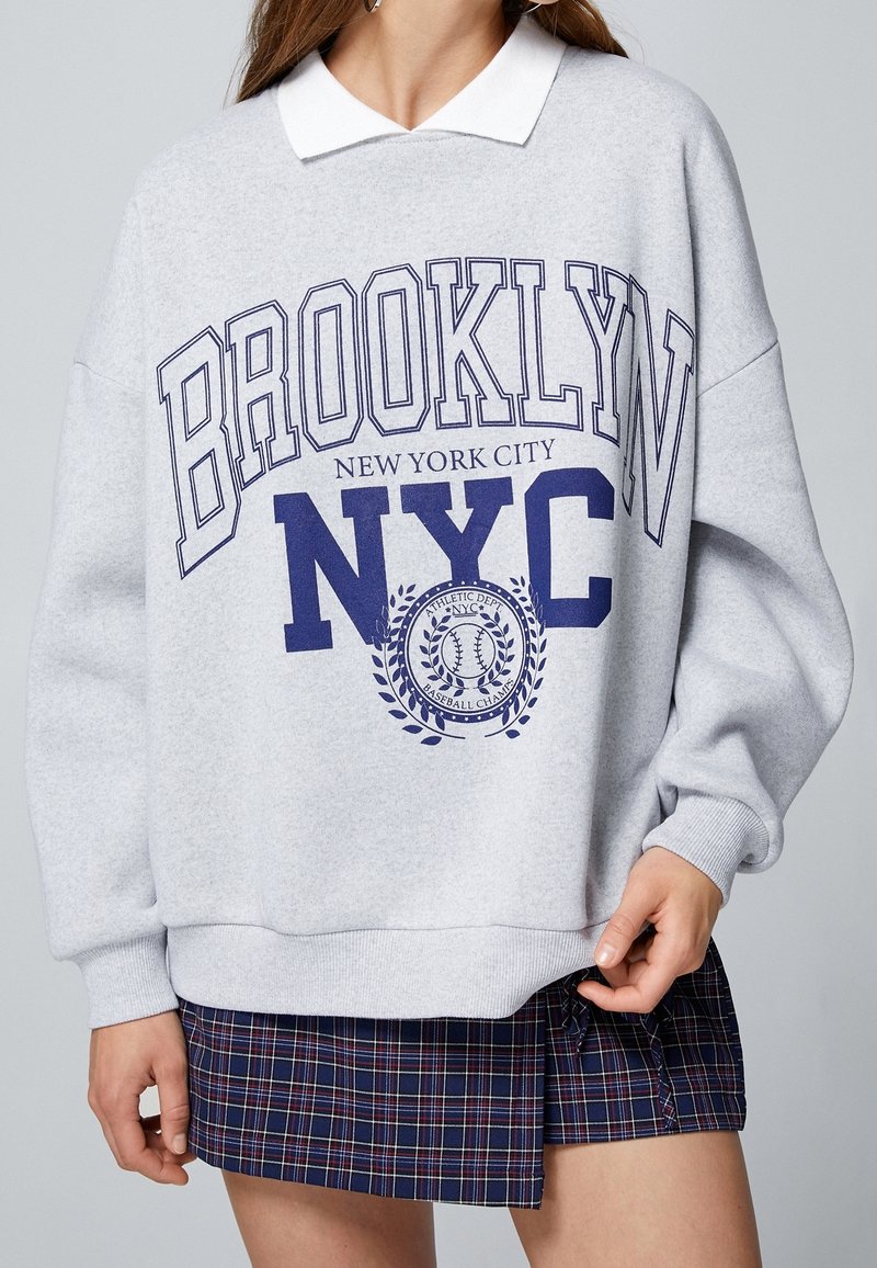 Femme portant un sweat-shirt gris Brooklyn NYC avec un col blanc et une jupe à carreaux bleue, la main reposant à ses côtés.