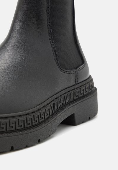 Botín negro de cuero con paneles laterales elásticos y suela de goma texturizada. Presenta un logo grabado a lo largo de la suela y un diseño elegante.