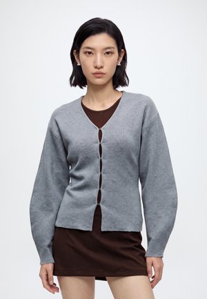 Donna con capelli corti neri che indossa orecchini di perle, un cardigan grigio abbottonato sopra un vestito marrone, in piedi su uno sfondo bianco semplice.