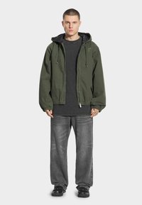 Zielona oliwkowa kurtka zapinana na zamek z kapturem, zestawiona z czarnym T-shirtem i szarymi dżinsami o luźnym kroju. Posiada elastyczne mankiety i nowoczesne sneakersy.