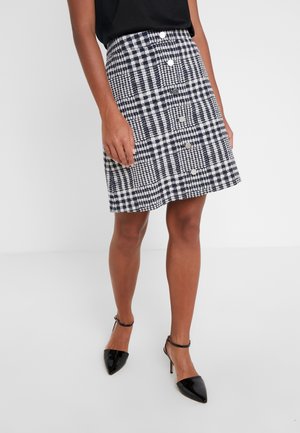 Femme portant une jupe à carreaux noir et blanc boutonnée sur le devant, un haut noir et des escarpins noirs à bride à la cheville sur fond blanc.