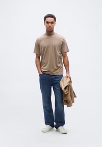 Μπεζ κοντομάνικη μπλούζα με ψηλό γιακά, τζιν παντελόνι και ανοιχτόχρωμα αθλητικά παπούτσια. Κρατάει ένα ανοιχτό καφέ σακάκι. Απλή, άνετη εφαρμογή.