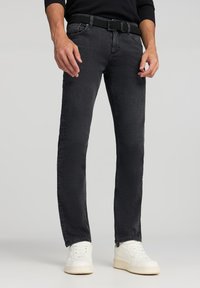 Jeans in denim nero con taglio dritto, caratterizzati da una cintura tessuta testurizzata. Il modello indossa scarpe da ginnastica bianche. Lo sfondo è semplice.