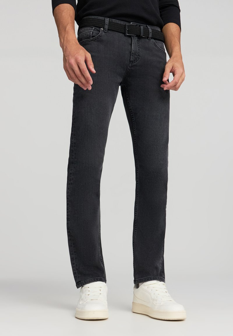 Jeans in denim nero con taglio dritto, caratterizzati da una cintura tessuta testurizzata. Il modello indossa scarpe da ginnastica bianche. Lo sfondo è semplice.