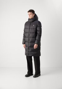 Schwarze Pufferjacke mit hohem Kragen und Kapuze, Reißverschluss und gesteppter Optik. Mit Branding auf der Brust und dem Ärmel. Kombiniert mit schwarzen Hosen.