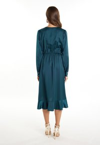 Femme debout de dos, portant une robe midi en satin teal à manches longues et à ourlet volanté, assortie de sandales à talons hauts dorées.