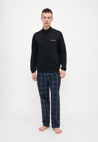 Black zip-up jacket with stand collar, paired with navy plaid pants featuring white lines. Model stands barefoot on a plain background.