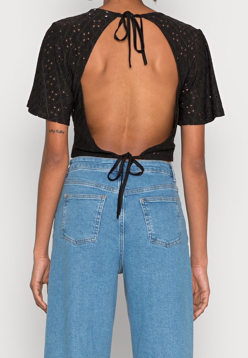 Top negro corto con mangas cortas y acampanadas, espalda descubierta atada con un lazo negro. Combinado con pantalones vaqueros de mezclilla azul claro de cintura alta.