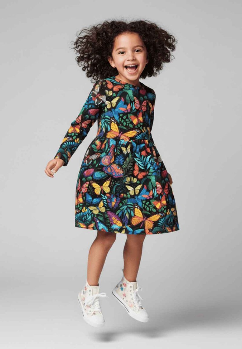 Kolorowa sukienka z długim rękawem z motywem motyli i liści na czarnym tle, w połączeniu z białymi wysokimi trampkami w kwiatowy wzór.