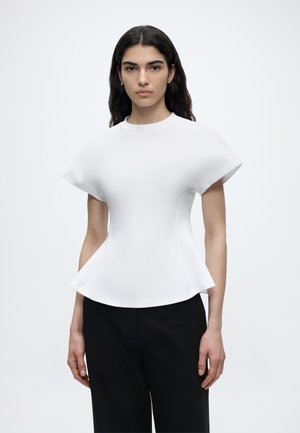 Frau mit langen dunklen Haaren, die ein weißes eng anliegendes Oberteil mit ausgestellten Ärmeln und Saum trägt, kombiniert mit schwarzen Hosen, stehend vor einem schlichten Hintergrund.