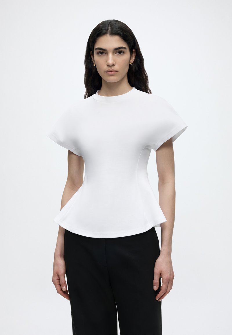 Γυναίκα με μακριά σκούρα μαλλιά που φοράει άσπρο εφαρμοστό τοπ με φουσκωτά μανίκια και τελείωμα, συνδυασμένο με μαύρο παντελόνι, στέκεται μπροστά σε απλό φόντο.