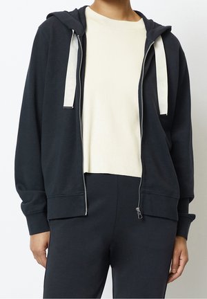 Black zip-up hoodie with a hood, white drawstrings, and ribbed cuffs. Layered over a cream ribbed top, paired with black pants.