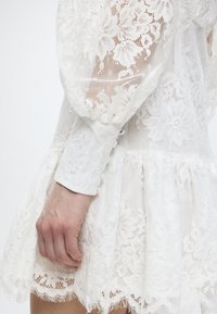 Χέρι που ακουμπάει δίπλα σε λευκό φόρεμα με δαντελένια λουλούδια, μακριά φουσκωτά μανίκια και λεπτομέρειες μανσέτας με κουμπιά.