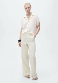 Cream short-sleeve button-up shirt and wide-leg cream trousers. Smooth fabric with a relaxed fit. Minimalist design; flat shoes and dark bracelet.
