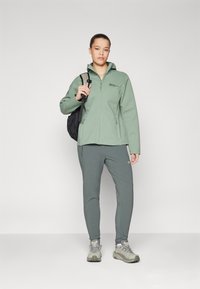 Grüne Fleecejacke mit Reißverschluss und Kapuze, kombiniert mit passenden Slim-Fit-Hosen und hellgrauen Wanderschuhen. Schwarzer Rucksack über der Schulter.