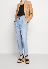 Hnědá kožená motocyklová bunda, černé tričko, světlé modré džíny s vysokým pasem a detaily na knoflících, a černé platformové sandály se dřevěnou podrážkou.