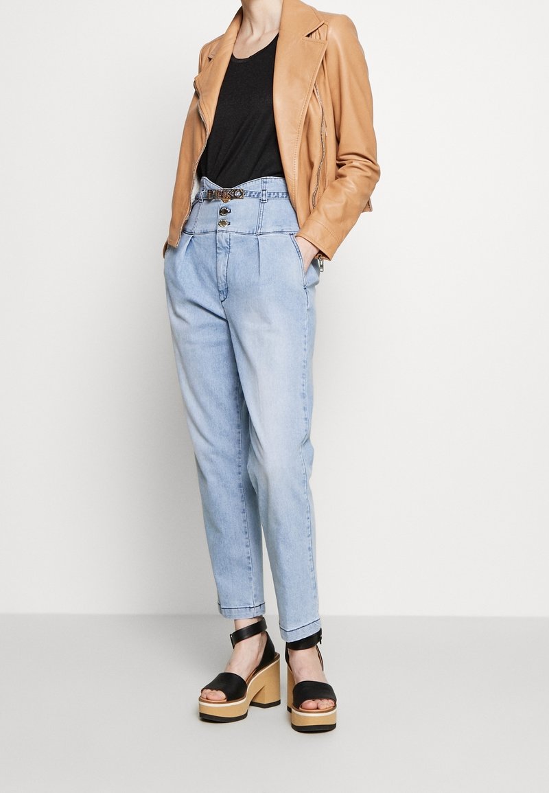 Hnědá kožená motocyklová bunda, černé tričko, světlé modré džíny s vysokým pasem a detaily na knoflících, a černé platformové sandály se dřevěnou podrážkou.