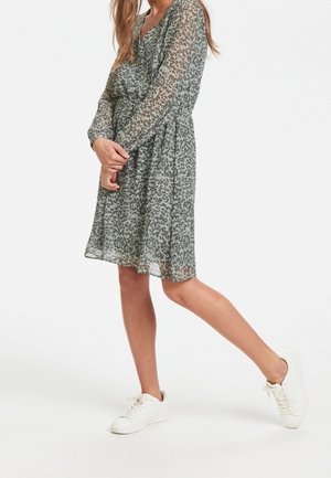 Robe verte à motifs avec des manches longues et une taille cintrée, en tissu léger ; assortie à des baskets blanches.