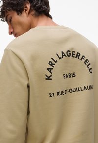 Khaki mikina s čiernym potlačeným textom na zadnej strane, ktorý znie: "KARL LAGERFELD PARIS 21 RUE ST-GUILLAUM!" Hladká textúra.