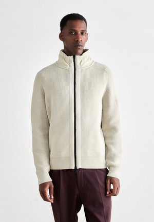Man wearing a cream ribbed zip-up sweater with a high collar and dark maroon trousers, standing against a plain white background.