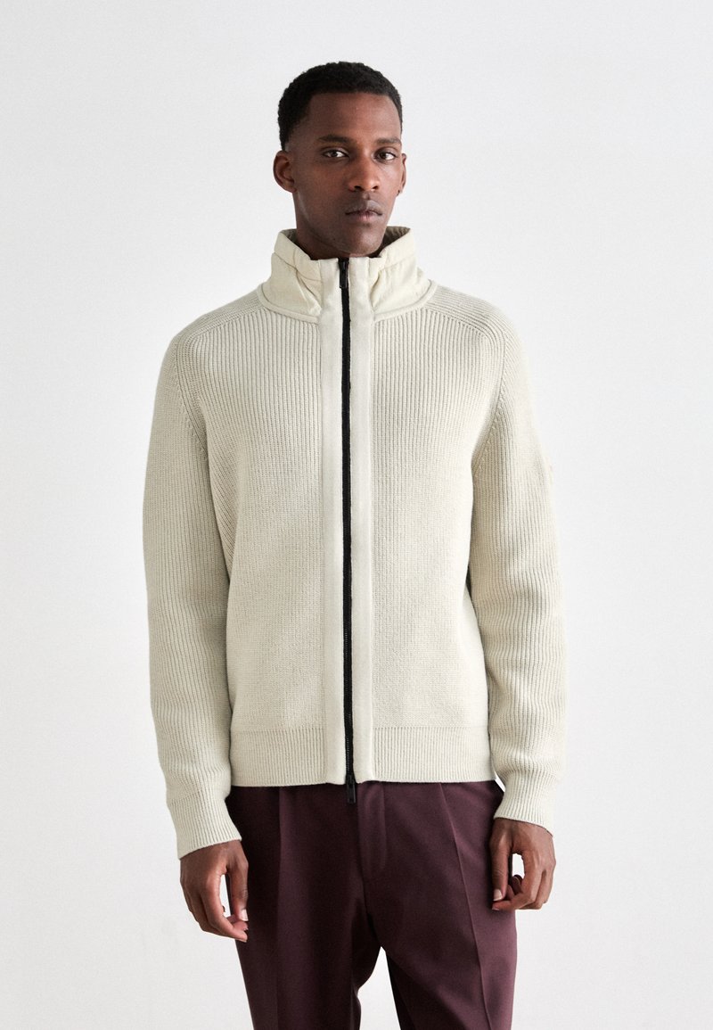 Homme portant un pull zippé côtelé crème avec un col haut et un pantalon bordeaux foncé, se tenant devant un fond blanc uni.