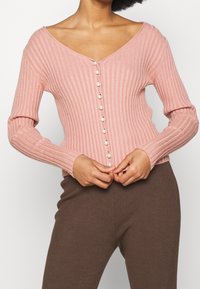 Jasnoróżowy prążkowany kardigan z długimi rękawami, dekoltem w serek oraz perełkowymi guzikami z przodu. Noszony w zestawie z brązowymi prążkowanymi spodniami.