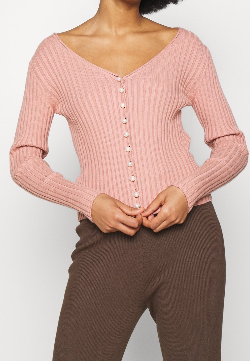 Jasnoróżowy prążkowany kardigan z długimi rękawami, dekoltem w serek oraz perełkowymi guzikami z przodu. Noszony w zestawie z brązowymi prążkowanymi spodniami.