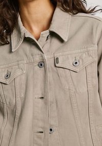 Chaqueta de mezclilla beige con cuello puntiagudo, bolsillos frontales con solapa y cierres de hardware plateado. Tejido texturizado con detalles de costura prominentes.