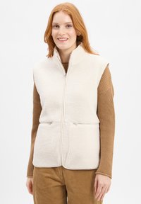 Fleece vest in off-wit met een hoge kraag, verticale ritszakken en een zachte, gestructureerde oppervlakte. Gemaakt over een bruine trui.