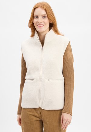 Fleece vest in off-white with a high collar, vertical zipper pockets, and a soft, textured surface. Worn over a brown sweater.