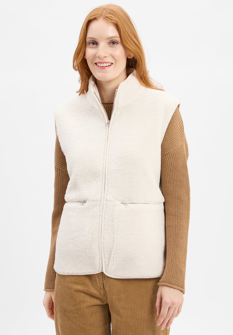 Fleece vest in off-wit met een hoge kraag, verticale ritszakken en een zachte, gestructureerde oppervlakte. Gemaakt over een bruine trui.