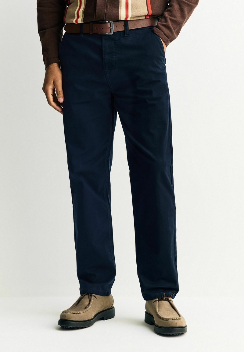 Tengerészkék vászon nadrág, pamut keverék anyagból készült, egyenes lábakkal, övvel és világosbarna cipővel párosítva.