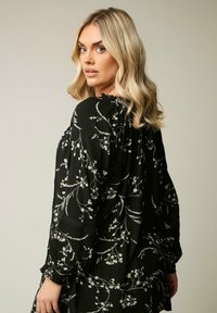 Blusa nera a maniche lunghe con motivi floreali bianchi, tessuto texturizzato, scollatura arricciata e dettagli in pizzo intricato sulle maniche.