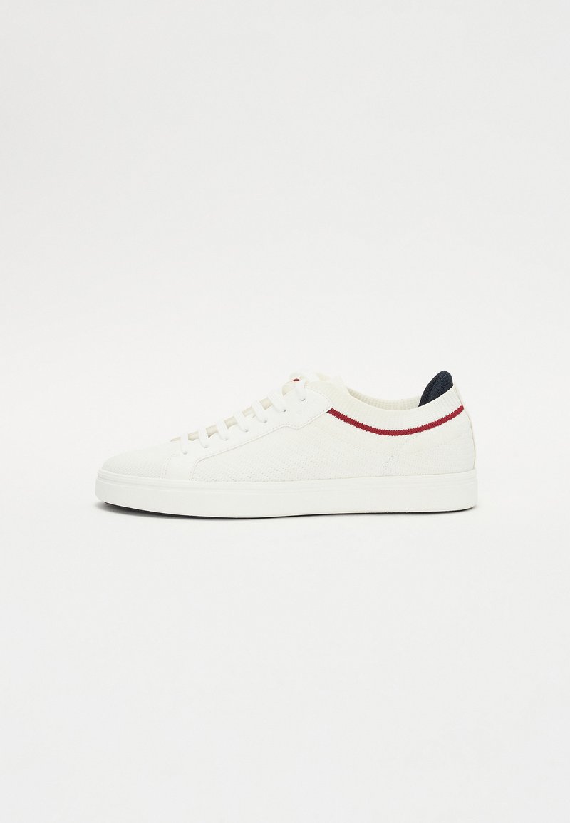 Hvit sneaker med teksturert strikket overdel, rød og blå aksentstriper, rund tå og flat gummisåle. Snøring for sikker passform.