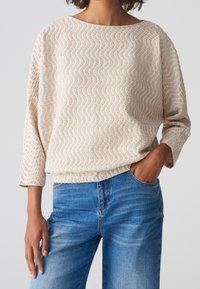 Pull beige texturé avec motif ondulé, col large et manches trois-quarts. Associé à un jean denim bleu avec une légère décoloration.
